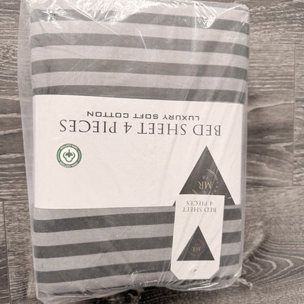 Packaged gray and white striped bed sheet set in clear plastic wrapping, with a white paper label reading 'BED SHEET 4 PIECES' and 'LUXURY SOFT COTTON', placed on a gray wood-textured surface.
