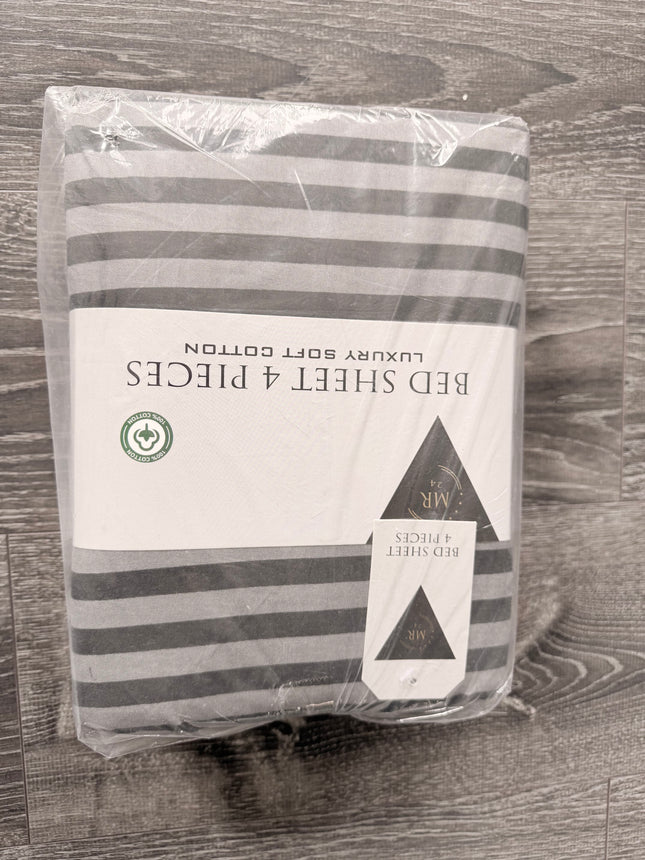 Packaged gray and white striped bed sheet set in clear plastic wrapping, with a white paper label reading 'BED SHEET 4 PIECES' and 'LUXURY SOFT COTTON', placed on a gray wood-textured surface.